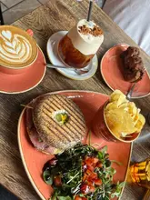A bright, natural-light flat lay of a brunch spread featuring a bagel, salad, chips, a latte, and an iced coffee. The image captures a casual, inviting cafe atmosphere.