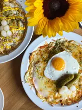 A bright, overhead flat-lay shot of two Swiss-style röstis served on white plates, garnished with pickles and a fried egg, accompanied by a large sunflower.