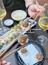 A high-quality, well-composed shot of a sushi spread at Sticks'n'Sushi, featuring a close-up of beef tataki on toast being held with chopsticks, with wine glasses and a blurred sushi platter in the background.