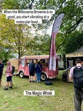 A candid shot of a pink food truck branded 'The Magic Whisk' situated at an outdoor food festival. The image captures the venue in its operational environment, surrounded by trees and other festival stalls, with customers visible in the foreground.