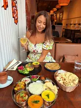 A bright, inviting photo of a woman posing at a table in an Indian restaurant, showcasing a generous spread of various dishes including a thali, rice, naan, and a glass of wine.