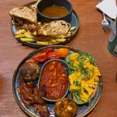 A high-quality, overhead shot of a brunch spread featuring a short rib grilled toastie with a side of jus and a 'big breakfast' plate containing eggs, bacon, sausage, beans, and a unique hash brown.