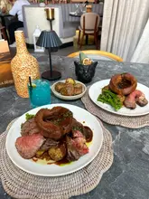 A high-quality, overhead shot of a Sunday roast meal at a restaurant, featuring two plates of beef roast with Yorkshire puddings, a blue cocktail, and a cocktail in a cat-shaped mug.