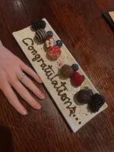 A high-quality, intimate shot capturing a celebratory dessert plate featuring chocolates and berries with 'Congratulations' written in chocolate sauce, accompanied by a hand displaying an engagement ring. The warm, ambient lighting and clean composition effectively convey a special, romantic occasion at a high-end venue.