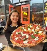 A high-quality, well-lit photograph featuring a smiling creator holding a large 18-inch pizza outside a restaurant. The pizza is clearly the focal point, showcasing fresh toppings like stracciatella and nduja, making it highly visually appealing.