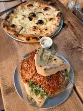 A high-quality, well-lit shot of a 'Vodka Parm Sando' and a pizza served on a rustic wooden table at a brewery taproom, effectively showcasing the casual dining experience.