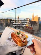 A high-quality, handheld shot of a lobster roll served on a rooftop terrace during golden hour. The image effectively captures the summer vibe, using the scenic city view as a backdrop to highlight the premium nature of the food.