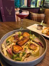A high-quality, well-composed shot of a scallop dish served at a Scottish restaurant, accompanied by a cocktail and a menu in the background, effectively capturing the dining atmosphere.
