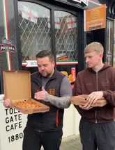 Two men stand outside a takeaway shop holding pizza boxes, likely filming a review or recommendation video for local food spots in the Bury and Bolton area.
