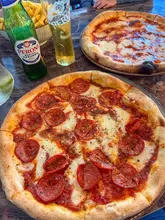 A casual, top-down shot of two pizzas, a portion of fries, and a glass of Peroni beer on a marble-effect table. The image captures a relaxed lunch setting, though the composition is slightly tight and includes an intrusive hand in the frame.