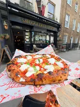 A high-quality, close-up shot of a burrata pizza held in front of the Smugglers Tavern, showcasing the fresh ingredients and charred crust. The image effectively captures the texture of the food and provides clear context of the venue.