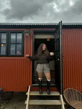A lifestyle shot featuring a person standing in the doorway of a shepherd's hut, holding a mug. The image captures a relaxed, countryside aesthetic, focusing on the accommodation and the 'slow living' vibe.