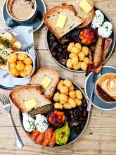 A high-quality, overhead flat-lay shot of a brunch spread featuring two main plates, two coffees, and a side of tater tots. The lighting is bright and natural, highlighting the textures of the sourdough, poached eggs, and various breakfast components.