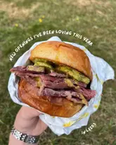 A close-up, handheld shot of a salt beef or pastrami sandwich in a paper wrapper, set against a grassy outdoor background, used to highlight a new food opening in Leeds.