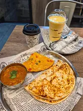 A top-down, well-lit shot of a Desi breakfast spread featuring a spicy omelette, a bowl of curry, flatbreads, a takeaway coffee cup, and a mango lassi, all served on branded newspaper-style paper.