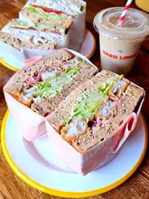 A high-quality, well-lit close-up shot of two generously filled sandwiches in paper wrappers, accompanied by an iced coffee. The focus is sharp on the textures of the bread, lettuce, and fillings, making for an appealing and casual lunch scene.