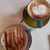 A top-down shot of two coffee cups, one featuring snail latte art, served on colourful ceramic saucers. The image captures a casual coffee break, highlighting the presentation of the drinks.