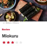 A professionally shot, overhead food photography image featuring a selection of Japanese dishes, including sushi rolls, served on minimalist, textured tableware. The composition is clean and highlights the freshness and presentation of the food, typical of a high-end restaurant review.