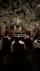 A short, romantic-themed reel showcasing the interior and food at Clos Maggiore in London. The video features shots of the flower-covered ceiling, the fireplace, and various dishes including steak tartare and fillet steak, presented as a recommendation for a date night venue.