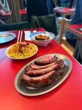A top-down, slightly angled shot of a BBQ meal featuring pork ribs, mac and cheese, and pork belly, served on a bright red table in a restaurant setting.