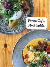 A top-down flat lay photograph of two brunch dishes served on a wooden table at Force Cafe in Ambleside. The image features a birria eggs benedict and a halloumi stack, showcasing fresh ingredients and vibrant colours.