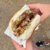 A close-up, handheld shot of a hog roast sandwich at a food market, showcasing the tender pork, stuffing, and crackling in a soft roll.