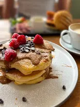 A close-up, top-down angled shot of a stack of vegan chocolate chip pancakes topped with fresh raspberries and biscoff sauce, with a coffee cup and a blurred burger in the background.