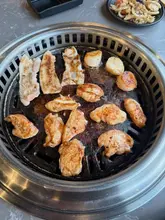 An overhead shot of a tabletop BBQ grill featuring various meats and scallops, likely at a Japanese Izakaya-style restaurant. The image captures the interactive cooking experience but suffers from a messy presentation.