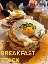 A close-up shot of a breakfast pancake stack topped with a fried egg, served at a cafe. The image captures the texture of the pancakes and the runny yolk, presented in a casual dining setting.
