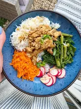 A vibrant, overhead shot of a homemade chicken teriyaki bowl featuring shredded chicken, rice, shredded carrots, green beans, kale, and sliced radishes, presented in a blue ceramic bowl.