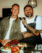 A candid, flash-lit portrait of two men posing at a table in a restaurant, with burgers and fried chicken in the foreground.
