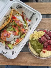 A bright, overhead shot of a breakfast spread featuring a stuffed pita wrap and a fruit bowl topped with white chocolate and pistachio cream, served in takeaway containers on a wooden table.