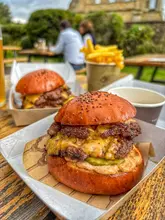 A high-quality, appetizing shot of a smash burger at The Abbey Inn. The natural lighting and shallow depth of field effectively highlight the texture of the burger, making it the clear focal point. The casual, outdoor setting is captured well, conveying a relaxed, enjoyable dining experience.
