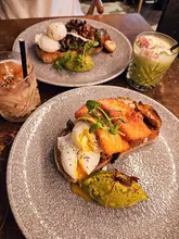 A top-down, overhead shot of a brunch spread featuring two plates of food and two drinks. The main focus is on a plate with poached eggs, halloumi, avocado, and mushrooms, while a second plate is partially visible at the top of the frame.