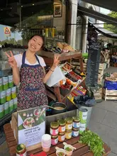 A friendly, authentic photo of a business owner at a market stall promoting Rempapa Spice Co., featuring jars of curry paste, fresh cilantro, and samples of curry.