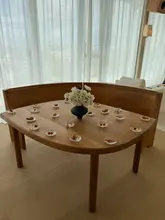 A minimalist, high-end restaurant interior shot featuring a wooden table set with sparse canapés and a central floral arrangement, bathed in bright natural light from a large window.