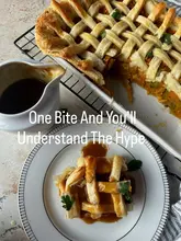 A well-composed, overhead shot of a homemade samosa pie featuring a golden, lattice-patterned puff pastry crust, served with a side of dark gravy. The lighting is soft and even, highlighting the texture of the pastry and the richness of the filling.