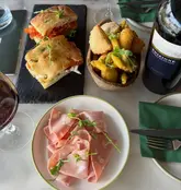 A high-quality, overhead flat-lay shot of an Italian meal featuring sliced mortadella, a fried appetizer platter, and a focaccia sandwich, accompanied by a bottle of red wine on a marble table.