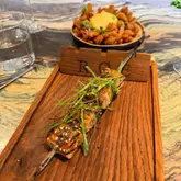 A high-quality, well-composed shot of a chicken tsukune skewer at Roe in Canary Wharf, presented on a branded wooden board with a blooming onion in the background.