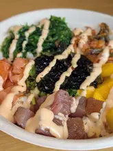 A close-up, top-down shot of a vibrant poké bowl featuring salmon, tuna, seaweed salad, kimchi, and mango, drizzled with spicy mayo. The image effectively showcases the freshness and variety of the ingredients, making it a classic, appealing food-focused shot suitable for social media promotion.