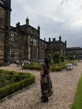 A travel-focused shot featuring a guest standing in the courtyard of Mar Hall Resort in Scotland. The image captures the historic architecture of the venue against an overcast sky, highlighting the resort's grounds and outdoor seating area.