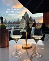A high-quality, atmospheric shot of two wine glasses on a table at a rooftop restaurant in Leeds, featuring a stunning sunset view of the city skyline.