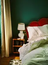 A moody, film-style photograph of a hotel bedroom interior, featuring a red velvet headboard against a dark green wall, capturing a relaxed and intimate atmosphere.