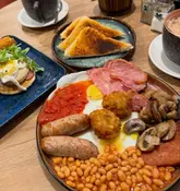 A top-down shot of a hearty cafe breakfast spread, featuring a full English breakfast with spam and a separate plate of pancakes with bacon and eggs.