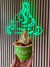 A casual, user-generated style photo of a frozen yogurt cup held in front of a neon sign at a Frurt venue.