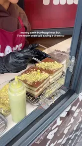 A short, static close-up video showing a gloved hand sprinkling crushed pistachios onto a square tiramisu dessert at a food stall.