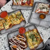 A top-down, overhead shot of four rectangular pizzas or flatbreads served on wire cooling racks on a speckled table, accompanied by small pots of dipping sauce.