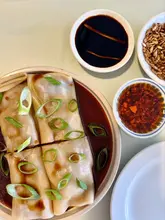 A clean, overhead shot of a dim sum dish, specifically beef cheung fun, accompanied by small bowls of chili oil and crispy toppings, presented on a light-colored table.