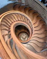 A high-angle, symmetrical shot looking down a striking wooden spiral staircase at a high-end restaurant. The image focuses on the architectural detail and the central light fixture, creating a visually compelling and sophisticated aesthetic.