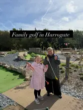 A bright, outdoor shot featuring a woman and a child posing with mini-golf clubs at a themed course in Harrogate. The image clearly communicates a family-friendly activity, with the text overlay providing context.